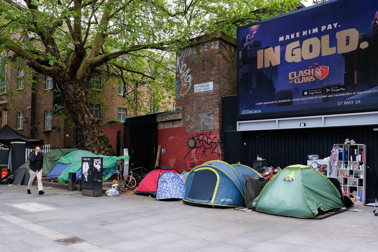 A person walks past a homeless encampment, seen with a row of tents under the backdrop of a noisy and busyTottenham Court Road.